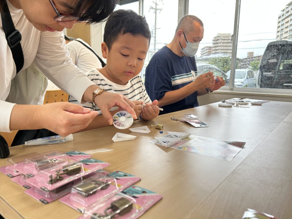 住宅展示場のイベントにて、小さなパーツを真剣に選ぶ男の子と、それを優しくサポートする親御様の様子。家族で協力してオリジナル南京錠をデコレーションする制作風景。