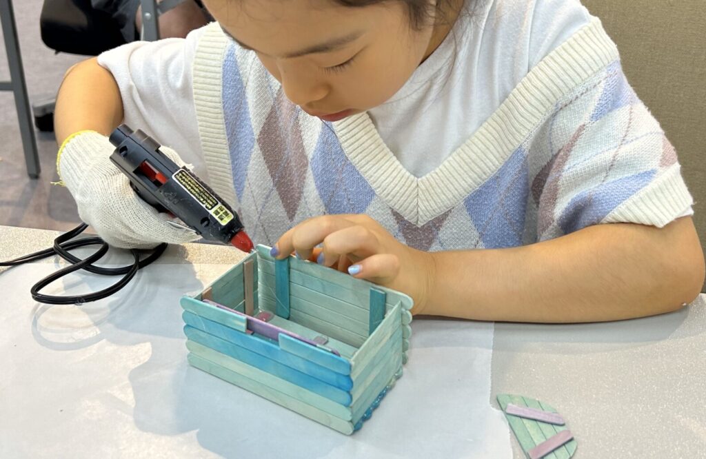 夏休みの工作イベントでアイスの棒を緻密に組み立てて作る立体的な貯金箱を子供がグルーガンを使用して制作する様子