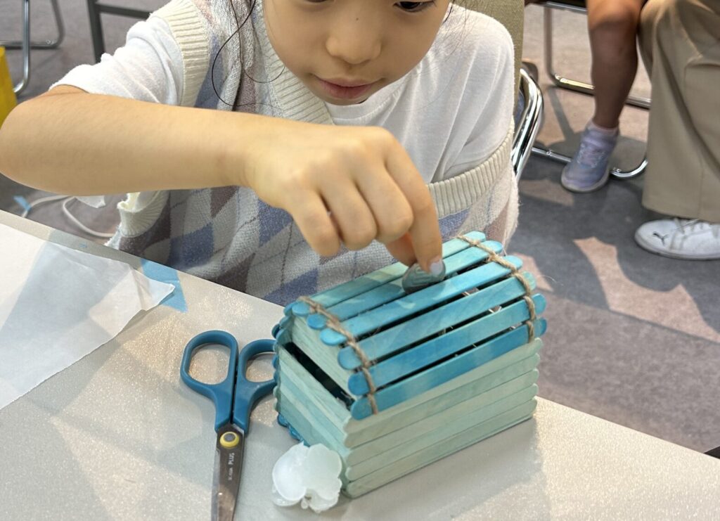 夏休みの工作イベントで子供がグルーガンを使って組み立てた貯金箱にコインが入るか自分で確認する様子